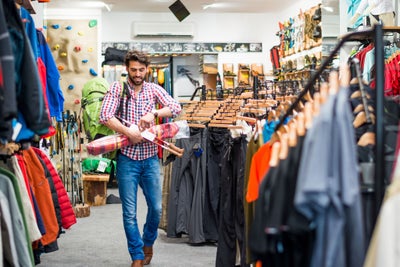 Bearded man in sports and outdoor equipment store looking to buy hiking equipment. He is holding green backpack, exercise mat, drinking bottle and hiking poles.